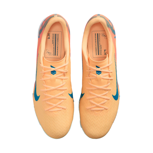 Top-down view of peach-colored Nike soccer cleats with blue accents and "KM" branding, isolated on a black background.