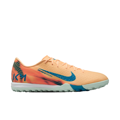 Side view of a peach-colored Nike Air Zoom soccer shoe with a blue swoosh, "KM" logo on the heel, and a mint green turf sole.