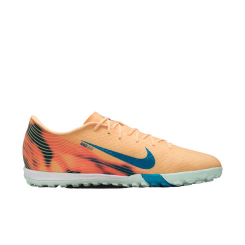 A peach-colored Nike turf soccer shoe featuring a blue swoosh, a graphic heel pattern, and a light green studded outsole.