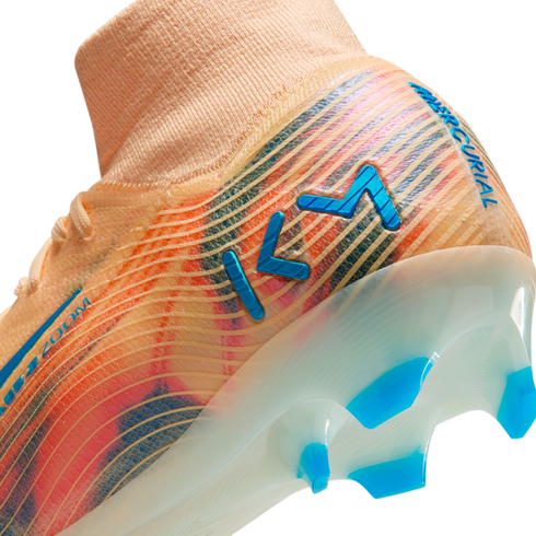 Close-up of a Nike Mercurial soccer cleat in orange with blue and pink accents, featuring a blue "KM" logo and a translucent sole with blue studs.