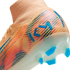 Close-up of a Nike Mercurial soccer cleat in orange with blue and pink accents, featuring a blue "KM" logo and a translucent sole with blue studs.