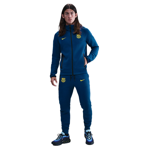 A young man with long hair stands in a blue FC Barcelona Nike tracksuit with yellow logos and blue sneakers against a black background.