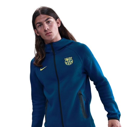 A young man with long brown hair wearing a blue Nike FC Barcelona zip-up hoodie against a black background.