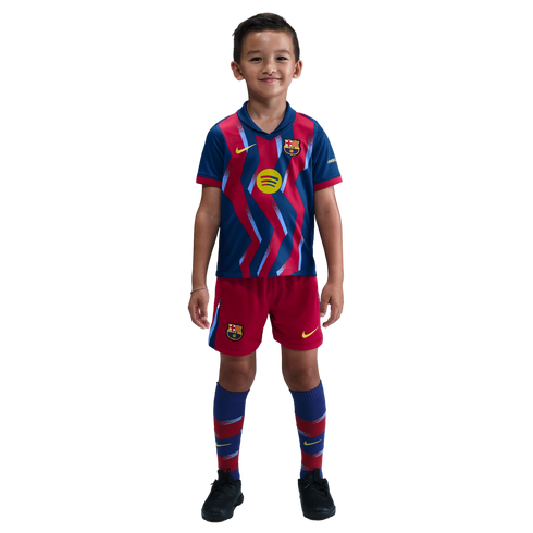 A young boy smiling in a blue and red zig-zag FC Barcelona soccer kit, including jersey, shorts, and socks, standing against a black background.