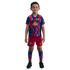 A young boy smiling in a blue and red zig-zag FC Barcelona soccer kit, including jersey, shorts, and socks, standing against a black background.