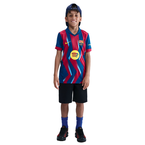 A smiling young boy wearing an FC Barcelona soccer jersey, black shorts, and a blue cap, standing against a solid black background.