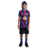 A smiling young boy wearing an FC Barcelona soccer jersey, black shorts, and a blue cap, standing against a solid black background.