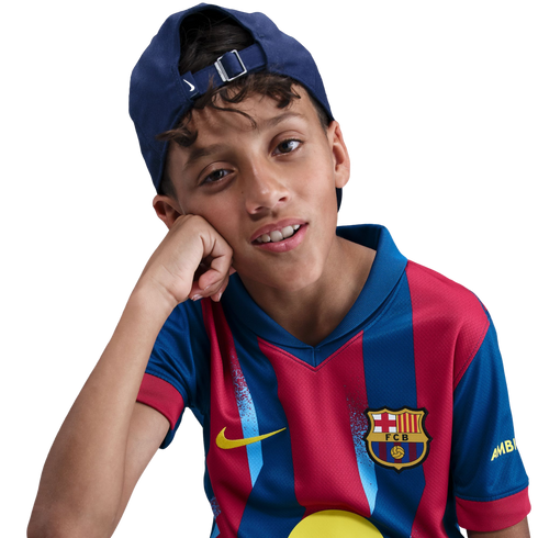 Portrait of a young boy wearing an FC Barcelona jersey and a backwards blue cap, resting his head on his hand against a black background.