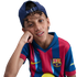 Portrait of a young boy wearing an FC Barcelona jersey and a backwards blue cap, resting his head on his hand against a black background.
