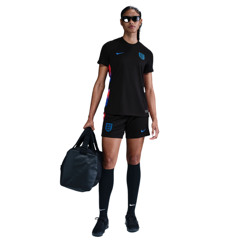 A woman standing in a black Nike England soccer kit and sunglasses, holding a black duffel bag.
