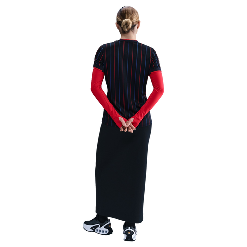 Back view of a person with a bun wearing a black pinstriped top over long red sleeves, a long black skirt, and black and white Nike sneakers.