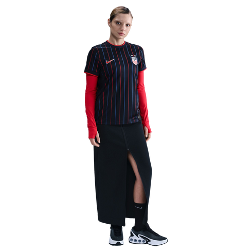 A woman in a striped black USA soccer jersey, red undershirt, long black skirt, and black sneakers, standing against a white background.