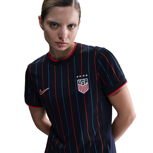 Woman wearing a black USA soccer jersey with red and blue pinstripes and four stars above the crest, posing against a white background.