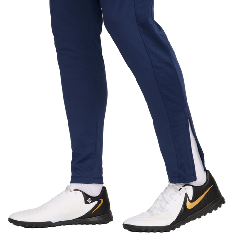 Side view of a person wearing navy blue track pants and white, black, and gold Nike turf shoes against a white background.