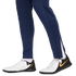 Side view of a person wearing navy blue track pants and white, black, and gold Nike turf shoes against a white background.