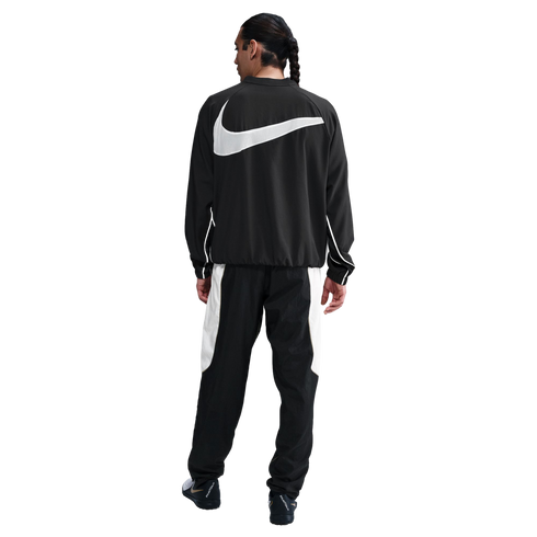 Back view of a man in a black Nike tracksuit featuring a large white swoosh on the jacket and white panels on the pants against a white background.