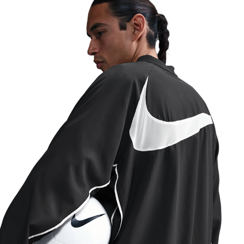 Man with a braid in a black Nike jacket with a large white swoosh on the back, holding a soccer ball. White background.
