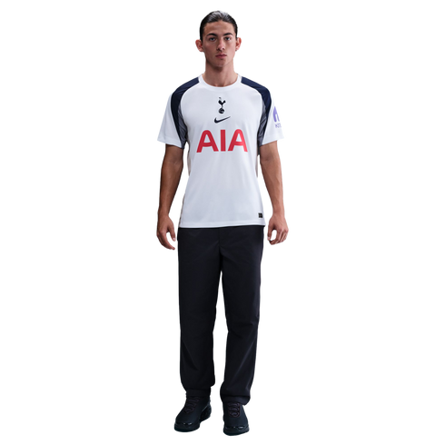 A man stands against a white background wearing a white and navy Tottenham Hotspur football shirt, black trousers, and black trainers.