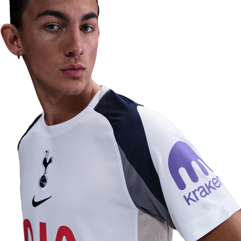 A man wearing a white Tottenham Hotspur jersey with the Kraken logo on the sleeve, looking to the side against a white background.