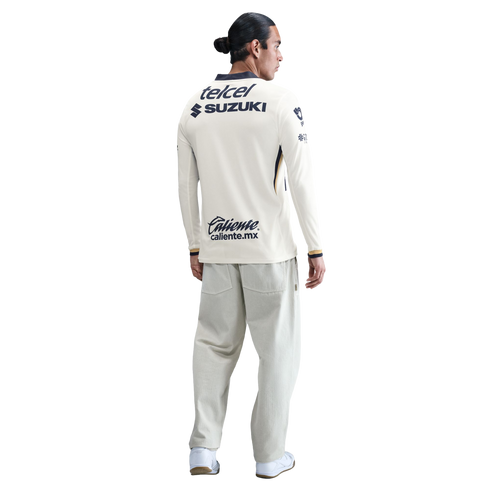 Back view of a man wearing a cream long-sleeved soccer jersey with sponsor logos and light gray wide-leg pants against a white background.