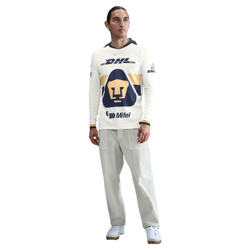 Man wearing a long-sleeved cream Pumas UNAM soccer jersey, light gray trousers, and white sneakers, standing against a plain white background.