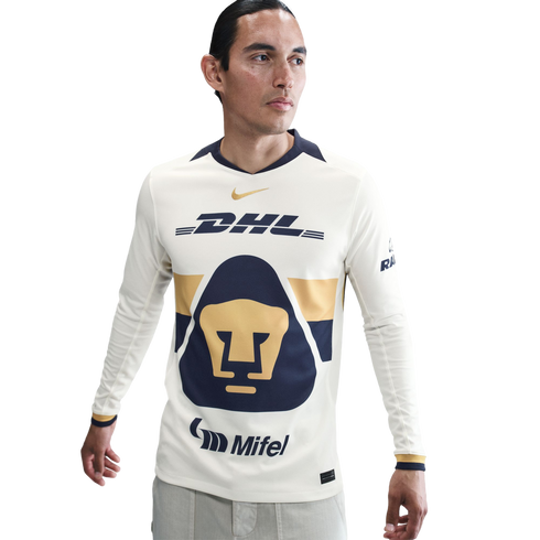 A man in a long-sleeved Pumas UNAM soccer jersey. The cream jersey features a large navy and gold feline head logo, with DHL, Nike, and Mifel branding.