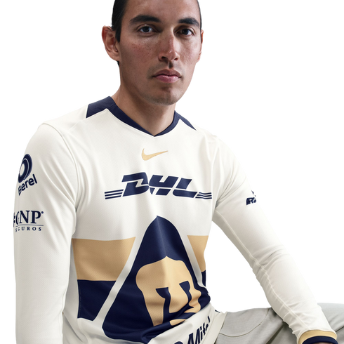A man in a cream-colored long-sleeve Pumas UNAM soccer jersey with navy and gold accents, featuring DHL and Nike logos, against a white background.
