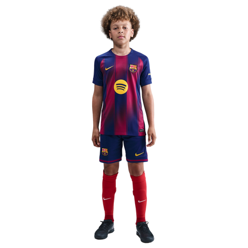 A young boy with curly hair stands against a black background wearing a full FC Barcelona football kit: blue and red striped jersey, blue shorts, and red socks.