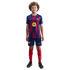A young boy with curly hair stands against a black background wearing a full FC Barcelona football kit: blue and red striped jersey, blue shorts, and red socks.