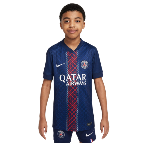 A young boy models a blue Paris Saint-Germain soccer jersey with a red central stripe and matching shorts against a white background. The kit features the club crest and Nike logo.