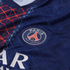Close-up of a blue Paris Saint-Germain soccer jersey featuring the embroidered team crest, red and white geometric patterns, and partial white sponsor lettering.