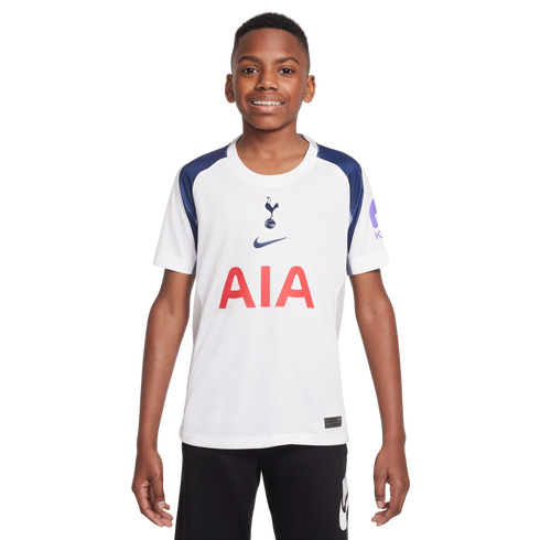 A smiling boy wears a white and navy Tottenham Hotspur soccer jersey with the red AIA logo and black shorts, standing against a white background.