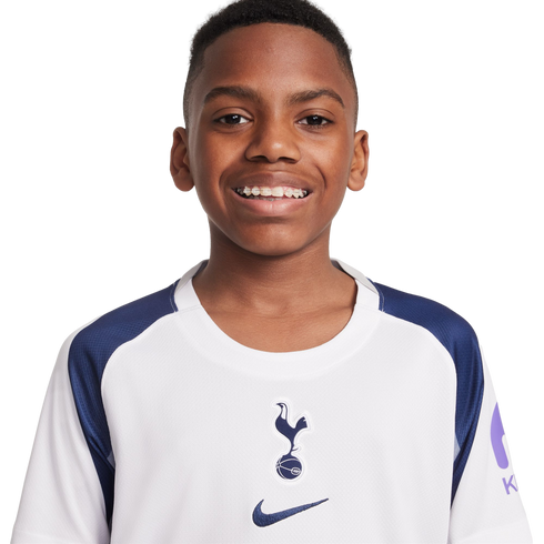 Portrait of a smiling young Black boy with braces, wearing a white and navy blue Tottenham Hotspur football jersey against a plain white background.