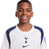 Portrait of a smiling young Black boy with braces, wearing a white and navy blue Tottenham Hotspur football jersey against a plain white background.