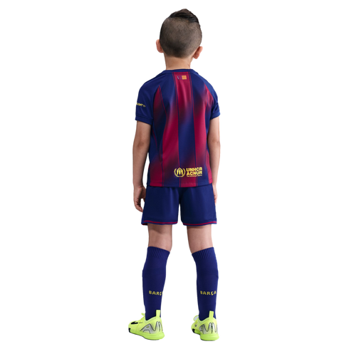 Back view of a boy in a blue and maroon FC Barcelona football kit and neon green cleats against a solid black background.