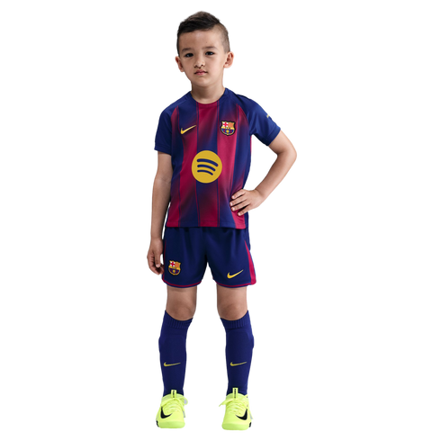 A young boy poses in a full FC Barcelona soccer kit with neon yellow cleats against a black background.