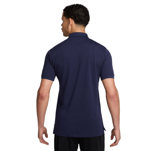 Rear view of a man wearing a navy blue short-sleeve polo shirt and black shorts against a solid black background.