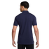Rear view of a man wearing a navy blue short-sleeve polo shirt and black shorts against a solid black background.