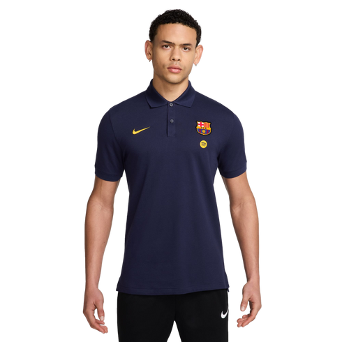 A man wearing a navy blue Nike FC Barcelona polo shirt with yellow branding and black Nike joggers, against a solid black background.
