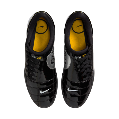 Top-down view of shiny black Nike Total 90 III soccer shoes with yellow insoles and small white swooshes on the toes, against a white background.