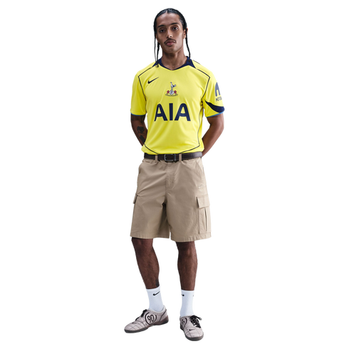 A man models a yellow Tottenham Hotspur football jersey, tan cargo shorts, and grey sneakers against a black background.