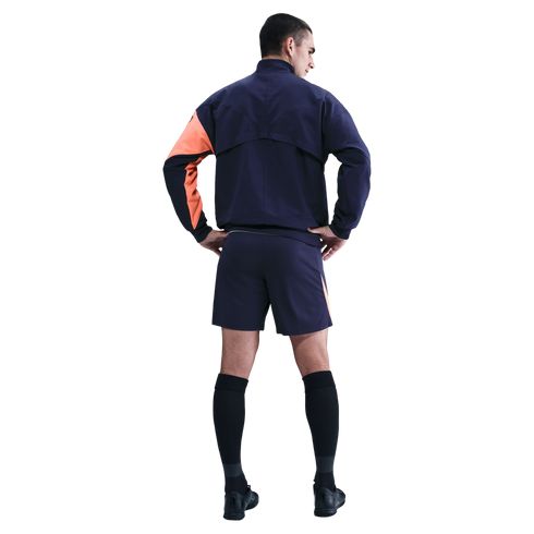 Back view of a man in a navy athletic jacket with one orange sleeve, navy shorts, black socks, and shoes, posing with hands on hips against a black background.