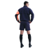 Back view of a man in a navy athletic jacket with one orange sleeve, navy shorts, black socks, and shoes, posing with hands on hips against a black background.