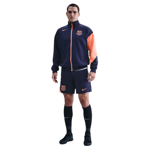 A man modeling a navy blue and orange Nike FC Barcelona track jacket and matching shorts against a solid black background.