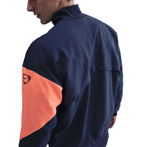 Side view of a person wearing a navy blue Nike jacket with a prominent orange sleeve panel against a black background.