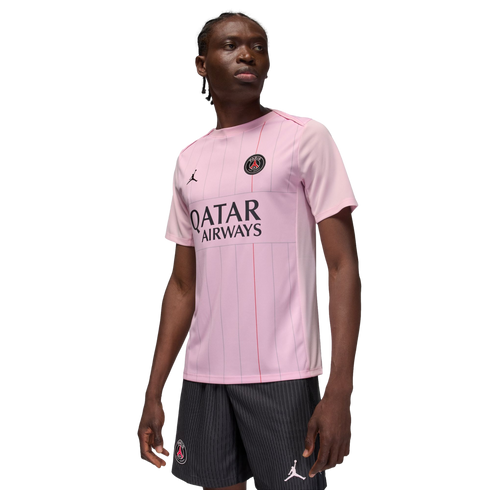 A man models a light pink Jordan Brand Paris Saint-Germain soccer jersey and matching black pinstripe shorts against a black background.