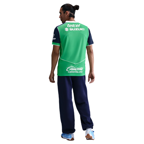 A man seen from behind wears a green and navy blue sports jersey with sponsor logos, navy pants, and light blue sneakers against a black background.