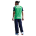 A man seen from behind wears a green and navy blue sports jersey with sponsor logos, navy pants, and light blue sneakers against a black background.
