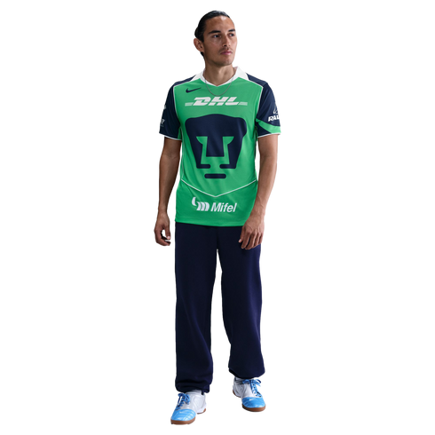 A man wearing a green and navy Pumas soccer jersey, navy sweatpants, and light blue sneakers stands against a black background.