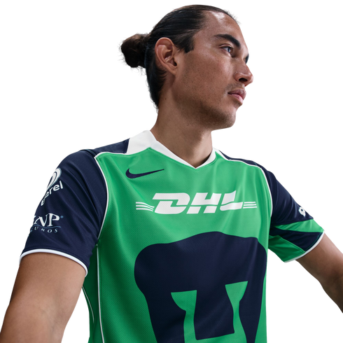 Man with hair in a bun wearing a green and navy Nike Pumas UNAM soccer jersey with a DHL logo, looking to the side.